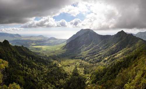 A panoramic view of lush green mountains and valleys under a cloudy sky, with the ocean visible in the distance.