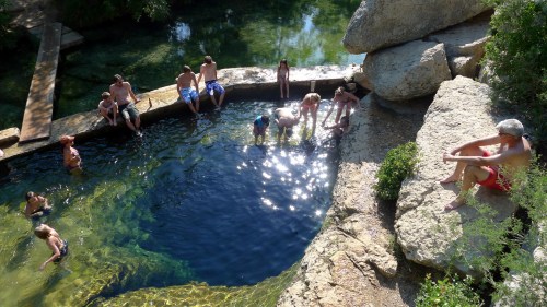 A group of people enjoying a sunny day by a natural swimming hole, with some sitting on rocks and others in the water.