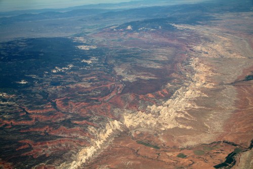 Aerial view of rugged, colorful terrain with layered rock formations and valleys under a clear sky.