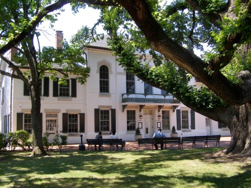 A historic white building with green shutters, surrounded by trees, with people sitting on benches in the foreground.