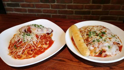 Two plates of pasta: one with fettuccine and marinara, topped with cheese, and another with chicken, cheese, and a breadstick.