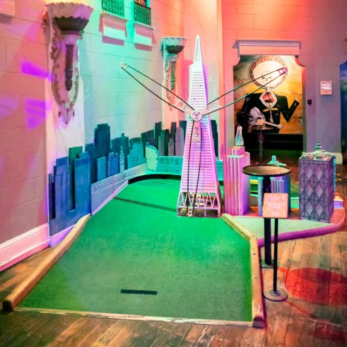 A colorful mini-golf course featuring a model of a skyscraper and city skyline, with vibrant lighting and decor.