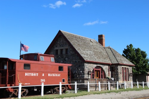 A red caboose labeled "Detroit & Mackinac" next to a stone train station under a blue sky with scattered clouds.