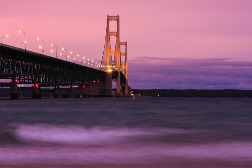 A bridge spans over water at sunset, with soft waves and a colorful sky in the background.