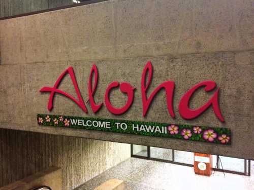 A colorful sign reading "Aloha" and "Welcome to Hawaii" with floral decorations on a textured wall.