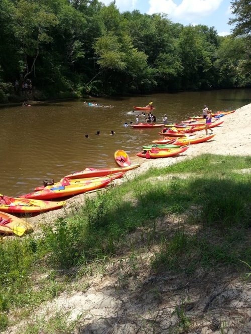 A sandy riverbank with colorful kayaks lined up, people swimming and enjoying a sunny day by the water.