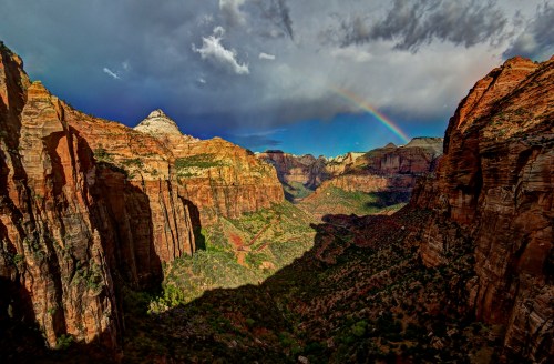 A stunning view of Zion Canyon with dramatic cliffs, a rainbow, and a moody sky after a rainstorm.