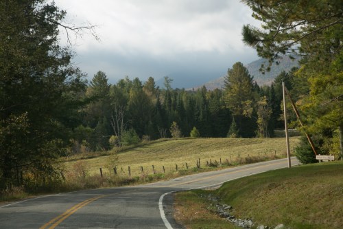 A winding road through a forested landscape with mountains in the background and a cloudy sky.