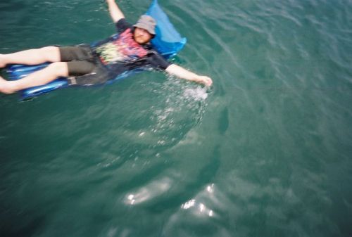 A person relaxes on a blue float in calm water, wearing a colorful shirt and a hat, enjoying a sunny day.