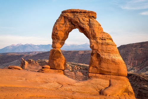 A natural rock arch stands tall against a backdrop of mountains and a colorful sky at sunset.