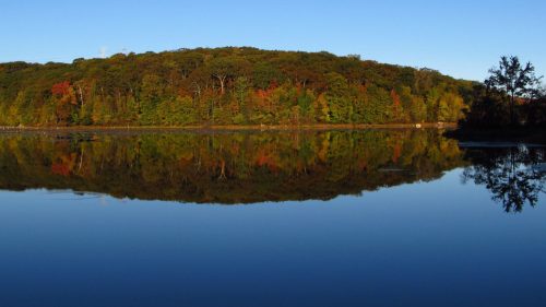 A serene lake reflecting a colorful forested hillside under a clear blue sky.