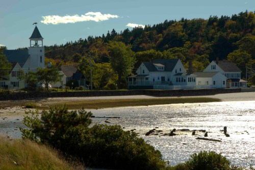 A serene coastal scene featuring a church, houses, and a calm waterway surrounded by trees and hills.