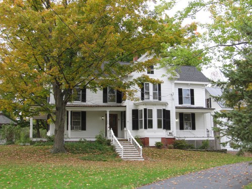 A large white house with black shutters, surrounded by trees and fallen leaves in autumn.