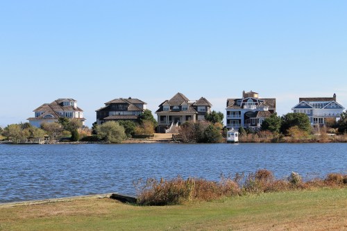 A row of waterfront houses with varied architecture beside a calm lake under a clear blue sky.