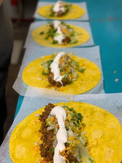 Four tacos on a blue table, filled with meat, lettuce, cheese, and topped with sour cream.