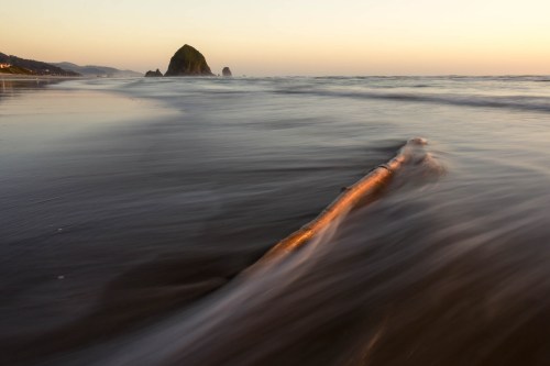 A serene beach scene at sunset, featuring gentle waves and a prominent rock formation in the distance.