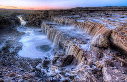 A stunning waterfall cascades over rocky cliffs, surrounded by a serene landscape at sunset.