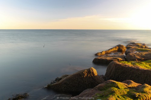 A serene seascape at sunset, featuring smooth water and rocky shoreline with green moss.