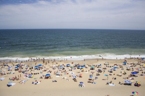 A crowded beach with colorful umbrellas, sunbathers, and gentle waves under a partly cloudy sky.
