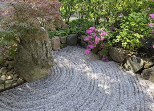 A serene garden scene featuring a raked gravel area, surrounded by rocks, vibrant flowers, and lush greenery.