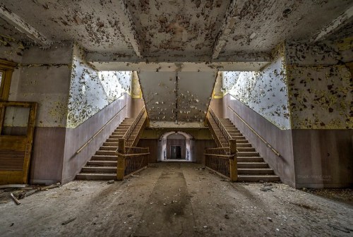 Abandoned interior of a building featuring two staircases, peeling walls, and scattered debris on the floor.