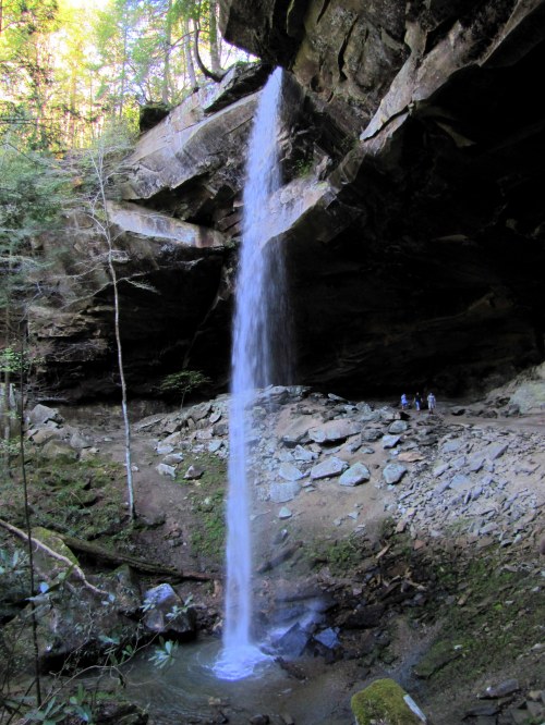 A waterfall cascades from a rocky overhang into a serene pool, surrounded by lush greenery and rocky terrain.