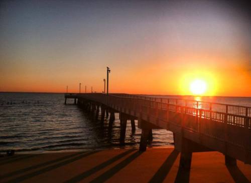 A serene sunset over a calm sea, with a pier extending into the water, casting long shadows on the sand.