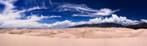 A panoramic view of vast sand dunes under a blue sky with wispy clouds and distant mountains.