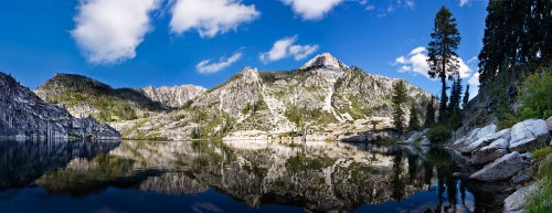 A serene mountain landscape reflecting in a calm lake, surrounded by trees and blue skies with scattered clouds.