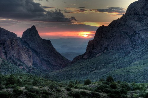 Sunset over rugged mountains, casting warm hues across a valley with lush greenery and dramatic rock formations.
