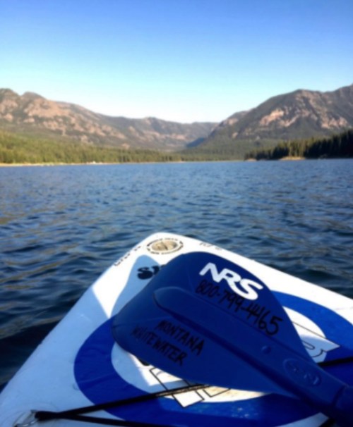 A paddle rests on a blue kayak, with mountains and a clear lake in the background under a bright blue sky.
