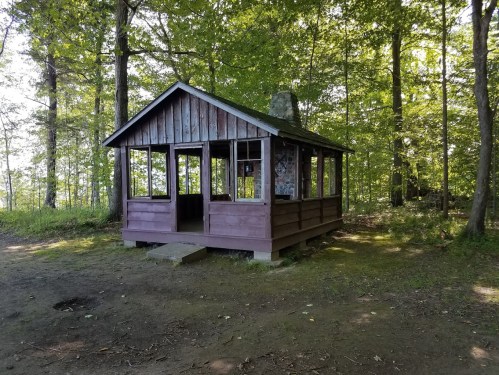 A small wooden cabin surrounded by trees, with a stone chimney and open windows, set in a natural forested area.