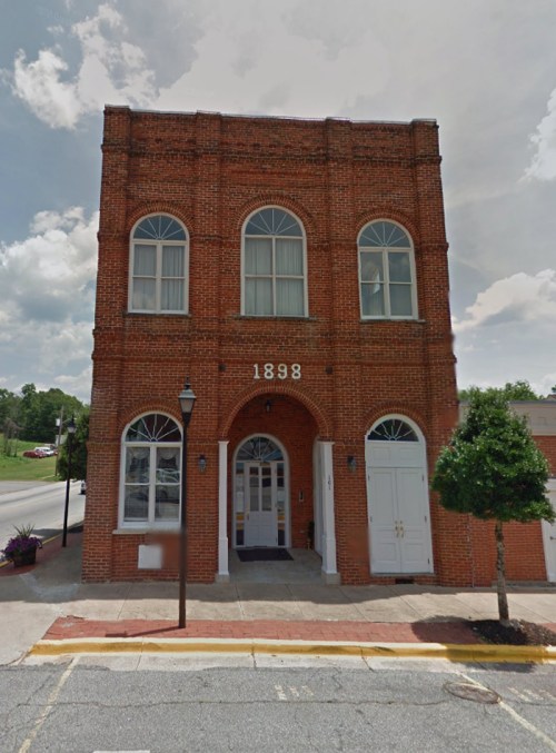 Historic brick building with large arched windows, featuring the year "1898" above the entrance, set against a cloudy sky.
