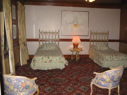 A cozy hotel room with two twin beds, floral bedding, a lamp, and vintage chairs on a patterned carpet.