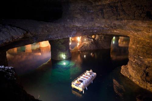 A small boat floats on calm water inside a dimly lit underground cavern with stone arches and reflections.