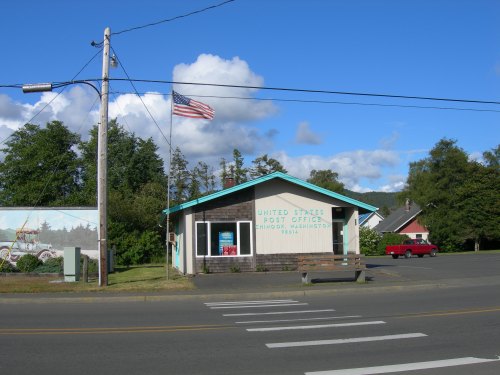 A small post office building with a flag, surrounded by trees and a clear blue sky. A red vehicle is parked nearby.