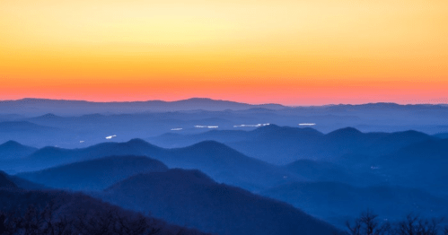 A serene mountain landscape at sunset, featuring layered hills and a gradient sky of orange and purple hues.