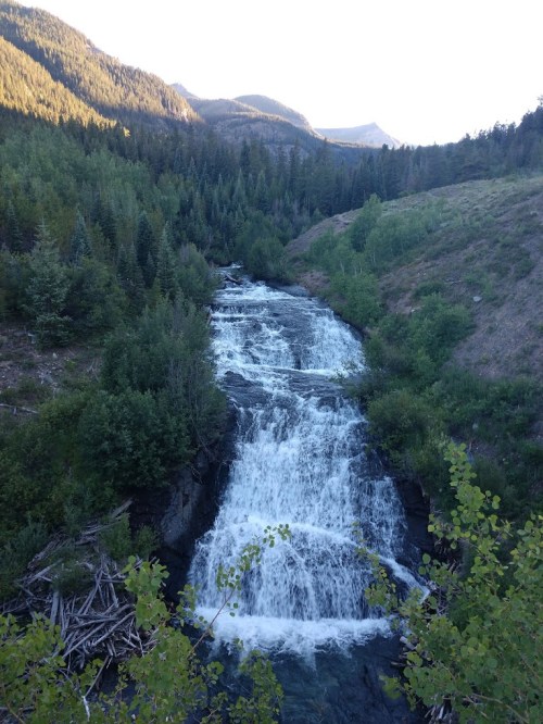 A cascading waterfall flows through a lush green landscape, surrounded by mountains and trees under a clear sky.