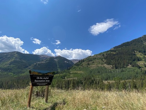 A sign for "Oh Be Joyfull Recreation Area" in a lush green valley with mountains and blue sky in the background.