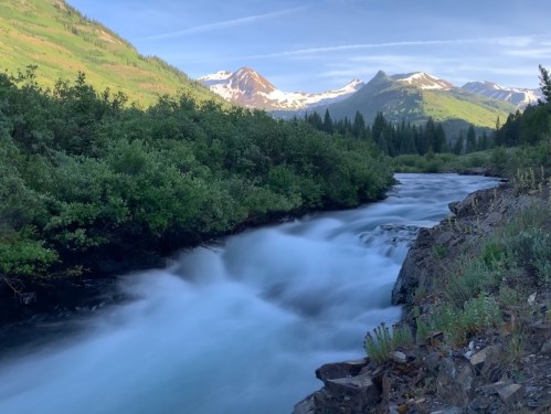 A serene river flows through lush greenery, with snow-capped mountains in the background under a clear blue sky.