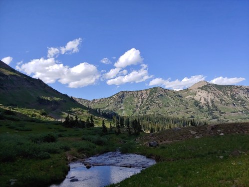 A serene landscape featuring a stream, green hills, and a blue sky with fluffy clouds.