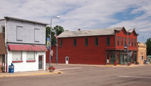 A small town intersection featuring a gray building, a red building, and a vintage storefront under a cloudy sky.