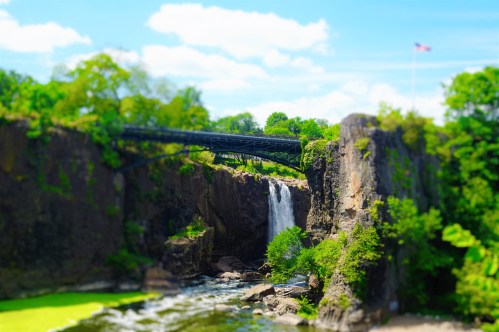 A scenic view of a waterfall surrounded by greenery, with a bridge overhead and a blue sky in the background.