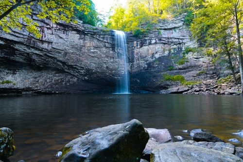 A serene waterfall cascades into a calm pool, surrounded by rocky cliffs and lush green trees.