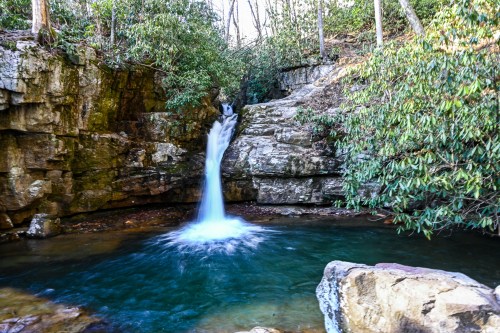 A serene waterfall cascades into a clear pool, surrounded by rocky cliffs and lush greenery.