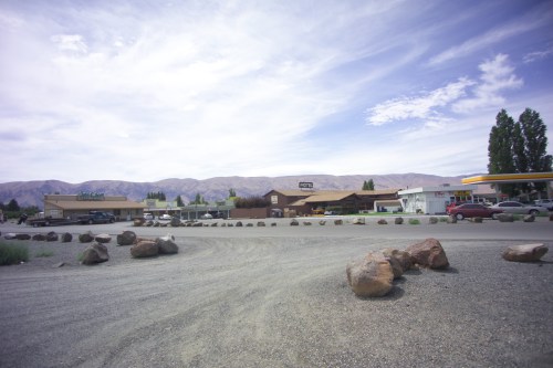 A gravel road leads to a small town with shops and a gas station, set against a backdrop of mountains and blue sky.