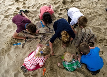 Children excavating a large fossil in the sand, using small tools and working together in a playful outdoor setting.