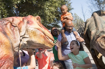 A group of children and adults interact with lifelike dinosaur models at an outdoor event, with excitement and joy.