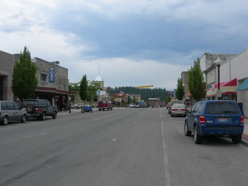 A quiet street in a small town, lined with trees and buildings, with parked cars and a cloudy sky above.