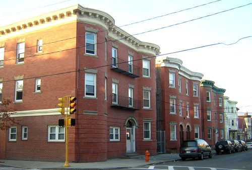 Red brick apartment building with curved edges, balconies, and a street corner with traffic lights and a fire hydrant.